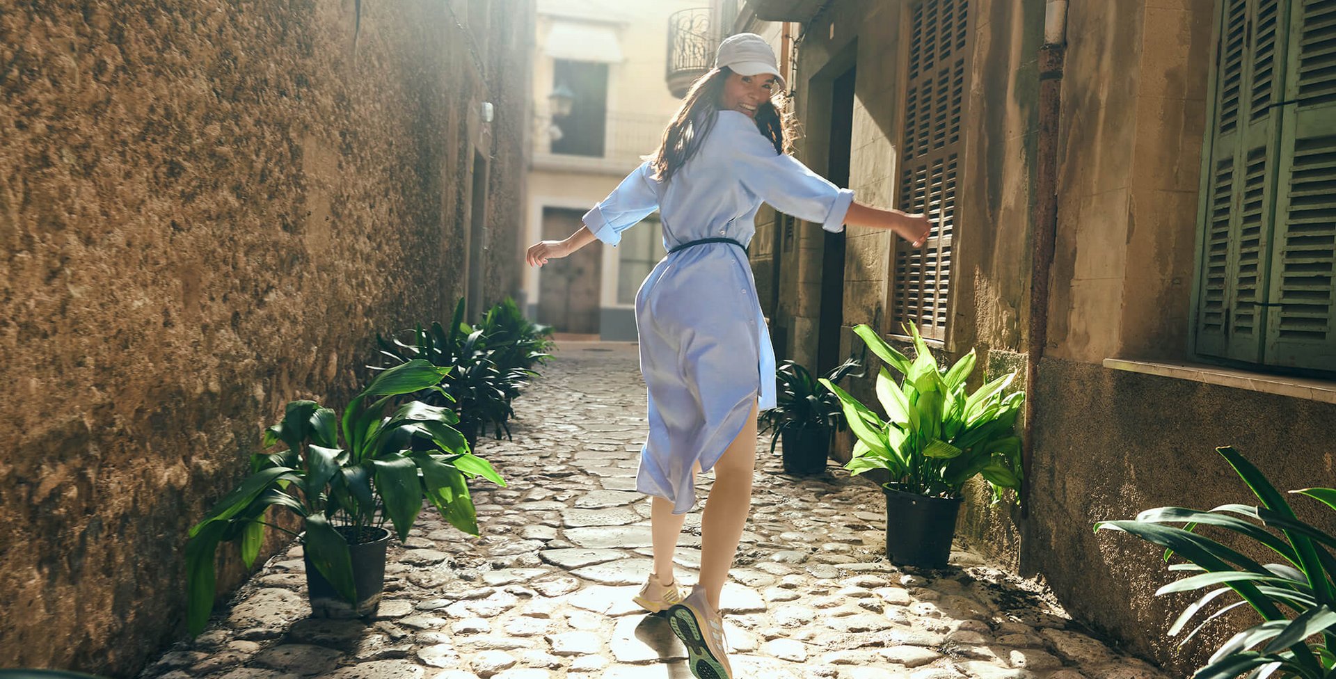 Eine junge Frau in einem hellblauen Kleid und Sneakers läuft fröhlich durch eine enge, mit Kopfsteinpflaster versehene Gasse. Die Sonne scheint durch die alten Gebäude und erzeugt ein warmes Licht. 